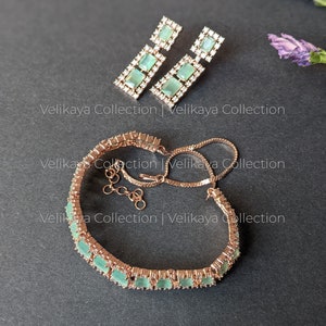 May include: A rose gold bracelet with a row of light green square-shaped stones set in a pave setting. The bracelet has a delicate chain and a lobster clasp. Matching earrings with a similar design are also pictured.