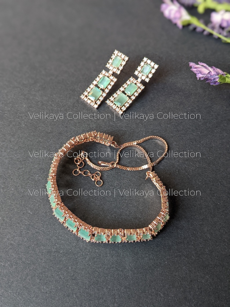 May include: A rose gold bracelet with a row of light green square-shaped stones set in a pave setting. The bracelet has a delicate chain and a lobster clasp. Matching earrings with a similar design are also pictured.