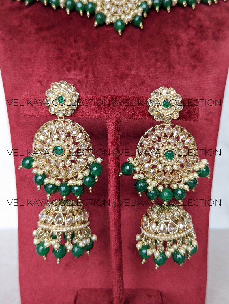 Puede incluir: Un conjunto de joyas de novia indias de oro y verde con un collar, pendientes y un maang tikka. El collar tiene un colgante redondo con dise&ntilde;os intrincados y cuentas verdes. Los pendientes tienen un dise&ntilde;o similar con un colgante redondo y cuentas verdes. El maang tikka tiene un peque&ntilde;o colgante con cuentas verdes.