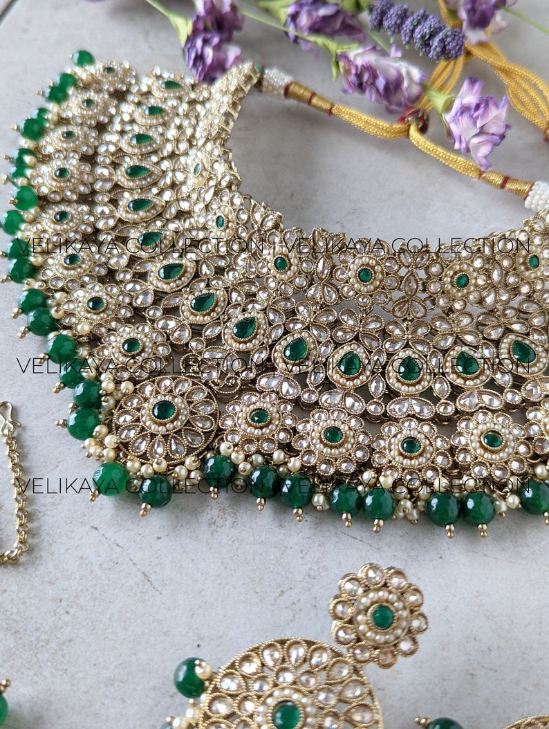 Puede incluir: Un conjunto de collar de novia indio de oro y verde con piedras verde esmeralda y perlas. El collar es de estilo gargantilla con un gran colgante en el centro. Los pendientes son peque&ntilde;os y redondos con un dise&ntilde;o similar.