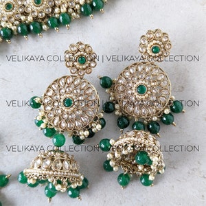 Puede incluir: Un conjunto de joyas de novia indias de oro y verde con un collar, pendientes y un maang tikka. El collar est&aacute; adornado con perlas y piedras verdes, y los pendientes son redondos con un dise&ntilde;o floral.