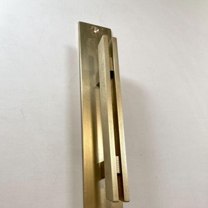 May include: A gold-toned metal door handle with a rectangular shape and a vertical, rectangular pull. The handle is mounted on a flat, rectangular plate.