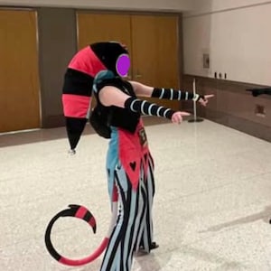 以下が含まれることがあります： 黒とピンクの道化師風の帽子、黒いベスト、青と黒のストライプのパンツで構成された衣装。お揃いのアームウォーマーと、ピンクのハートのディテールが付いた長く湾曲した黒とピンクの尻尾が含まれています。帽子には小さな鈴が付いています。
