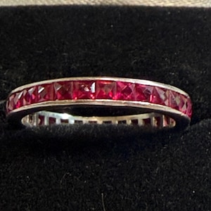 Puede incluir: Una alianza de tono plateado con una hilera continua de rubíes de color rojo intenso, tallados en forma cuadrada y engastados en canal. El anillo muestra una disposición uniforme de gemas facetadas que recorren toda la circunferencia.