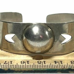 May include: A silver bracelet with a large, round, silver charm in the centre. The bracelet has a cutout design on either side of the charm.