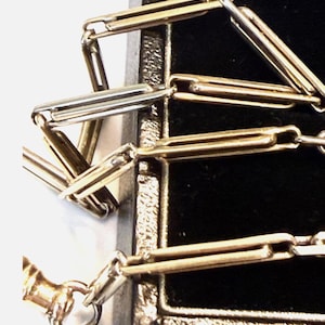 Puede incluir: Primer plano de un collar de cadena dorado y plateado. La cadena está compuesta por eslabones rectangulares, con un cierre visible en la parte inferior. El collar se muestra sobre un fondo negro.