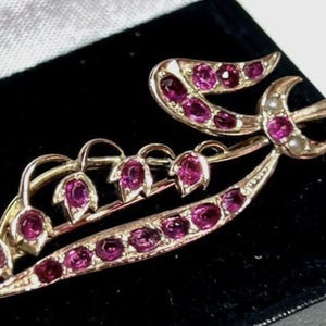 May include: A gold brooch featuring a lily of the valley design. The flowers are set with pink gemstones and the leaves are set with smaller pink gemstones.
