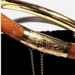 May include: A hinged bangle bracelet featuring a smooth, polished orange stone segment and a gold-toned metal section. The metal portion displays intricate engraved line patterns and a small heart-shaped detail, accompanied by a delicate safety chain.