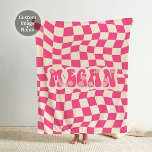 Puede incluir: Una manta a cuadros rosa y crema con el nombre "MEGAN" en una fuente retro. La manta tiene un patr&oacute;n de tablero de ajedrez ondulado y se muestra sobre un fondo neutro.