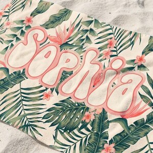 May include: A rectangular beach towel featuring a cream-colored background with a repeating pattern of green tropical palm leaves and small pink flowers. The name Sophia is printed across the center in large, bubble-style pink letters with a white outline.