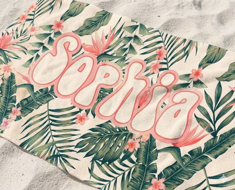 May include: A rectangular beach towel featuring a cream-colored background with a repeating pattern of green tropical palm leaves and small pink flowers. The name Sophia is printed across the center in large, bubble-style pink letters with a white outline.