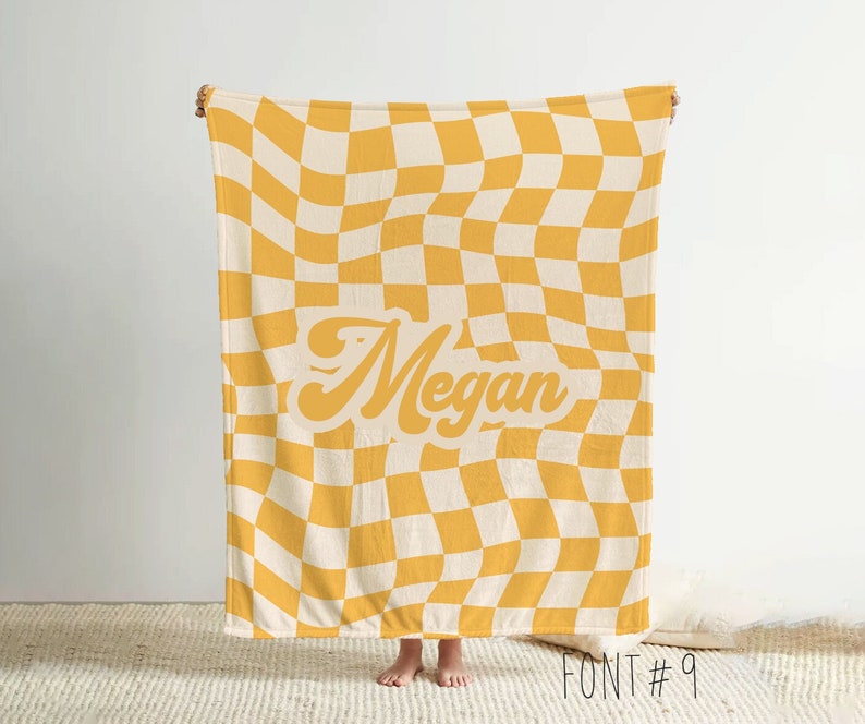 Puede incluir: Una manta a cuadros amarillos y crema con el nombre "Megan" en una fuente retro. La manta se sostiene, mostrando el patr&oacute;n completo y el texto. El texto "FONT #9" est&aacute; en la parte inferior derecha.