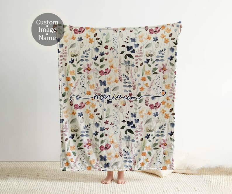 May include: A white fleece blanket with a floral pattern in shades of pink, blue, yellow, and green. The blanket has the name "mariarum" embroidered in blue cursive script.