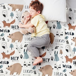 May include: A white baby blanket with a woodland animal print. The blanket features a repeating pattern of foxes, owls, deer, bears, and rabbits. The words "Hey Wild" and "Lucas" are repeated throughout the blanket.