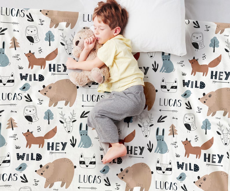 May include: A white baby blanket with a woodland animal print. The blanket features a repeating pattern of foxes, owls, deer, bears, and rabbits. The words "Hey Wild" and "Lucas" are repeated throughout the blanket.