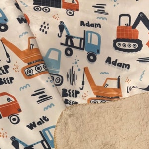 May include: A white fabric baby blanket with a colorful print of construction vehicles. The blanket features the names "Adam" and "Walt" printed on it. The blanket has a soft, fuzzy white backing.