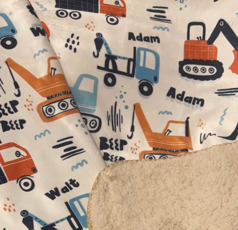 May include: A white fabric baby blanket with a colorful print of construction vehicles. The blanket features the names "Adam" and "Walt" printed on it. The blanket has a soft, fuzzy white backing.