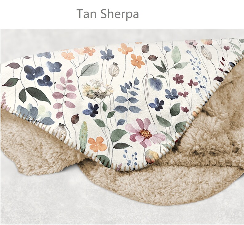 May include: Tan sherpa throw blanket with a floral print featuring watercolor flowers in shades of pink, blue, and orange on a white background. The blanket is folded in half, showing both the sherpa and floral sides.
