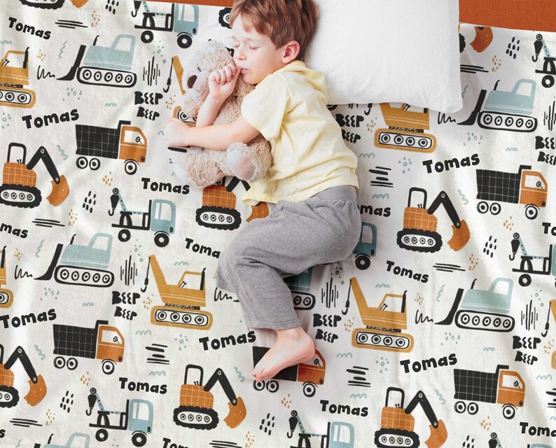 May include: A white baby blanket with a repeating pattern of construction vehicles in shades of blue, orange, and black. The vehicles include dump trucks, excavators, and tow trucks. The name "Tomas" is repeated throughout the blanket.