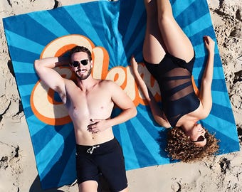 May include: A rectangular beach towel featuring a vibrant blue and light blue sunburst pattern with the word Aloha written in large, bold, orange and cream-colored retro-style lettering across the center of the fabric surface.