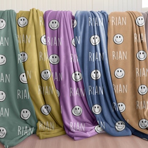 May include: Five fleece blankets with a repeating pattern of smiling faces and the name "RIAN" in white text. The blankets are in various colors: teal, yellow, purple, blue, and brown.