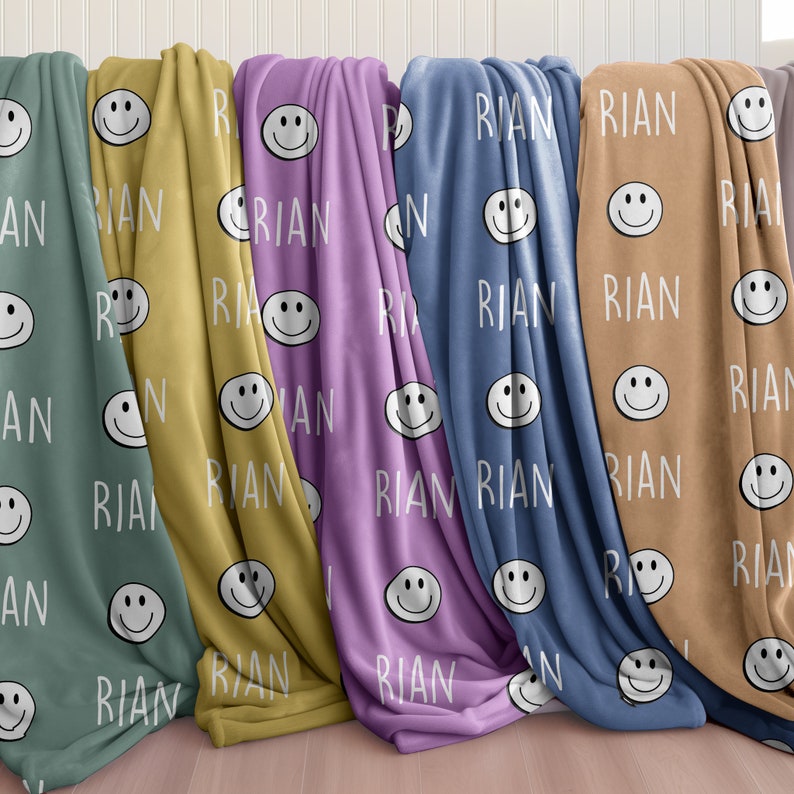 May include: Five fleece blankets with a repeating pattern of smiling faces and the name "RIAN" in white text. The blankets are in various colors: teal, yellow, purple, blue, and brown.
