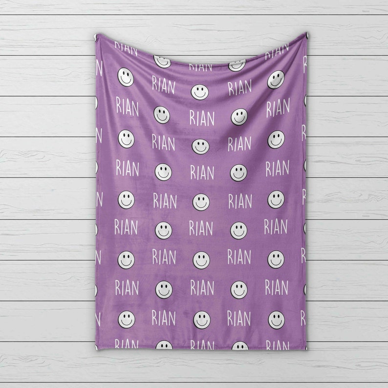 May include: Purple fleece blanket with a repeating pattern of white smiling faces and the name "RIAN" printed on it.