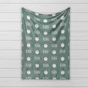 May include: A green fleece blanket with a repeating pattern of white smiling faces and the name "RIAN" printed on it.