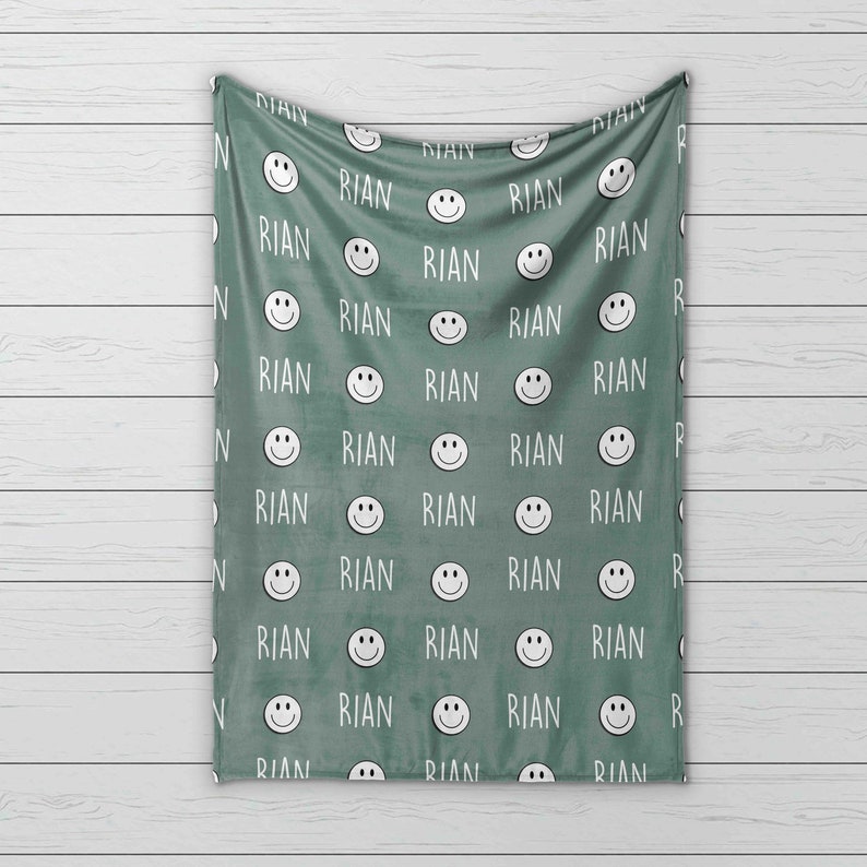 May include: A green fleece blanket with a repeating pattern of white smiling faces and the name "RIAN" printed on it.