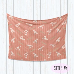 Puede incluir: Una manta de beb&eacute; rosa coral con un patr&oacute;n de lazos blancos y verdes y el nombre "Melanie" repetido en toda la superficie. La manta est&aacute; colgada sobre un fondo de madera blanca. STYLE #6