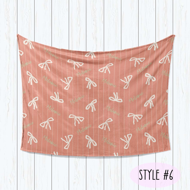 Puede incluir: Una manta de beb&eacute; rosa coral con un patr&oacute;n de lazos blancos y verdes y el nombre "Melanie" repetido en toda la superficie. La manta est&aacute; colgada sobre un fondo de madera blanca. STYLE #6