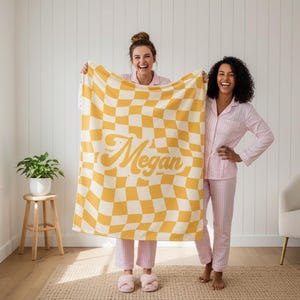 Puede incluir: Una manta a cuadros amarillos y blancos con el nombre "Megan" en una fuente retro. Dos personas con pijamas de rayas rosas sostienen la manta. La manta es un elemento de decoraci&oacute;n del hogar.