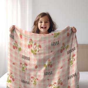 May include: A pink and white checkered baby blanket with a strawberry and flower pattern. The blanket has the name "HANNA" printed on it multiple times.