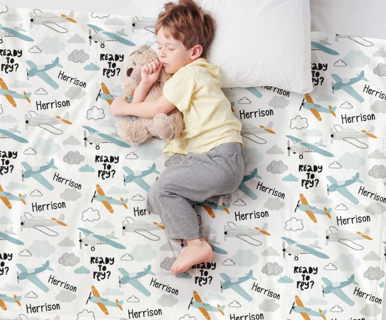 May include: A white baby blanket with a blue and orange airplane pattern. The blanket has the text "Ready to Fly?" repeated throughout. The name "Herrison" is also repeated throughout the blanket.
