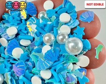 May include: A handful of blue and white craft embellishments. The mix includes seashell, star, and dolphin shapes, along with round and rod-shaped pieces. Large pearl beads are also present. The text "NOT EDIBLE" is visible.