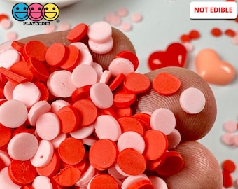 May include: A close-up image of red and pink round sprinkles. The sprinkles are scattered on a hand. The text "NOT EDIBLE" is visible in the upper right corner of the image.