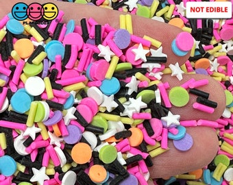 May include: A variety of colorful sprinkles, including stars, circles, and long, thin pieces. The sprinkles are in a hand, and the text "NOT EDIBLE" is visible in the upper right corner of the image.