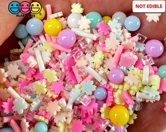 May include: A variety of pastel-colored sprinkles, including stars, flowers, and small balls, in a hand. The sprinkles are not edible. "NOT EDIBLE" is written in black text on a white background in the upper right corner of the image.