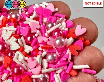 May include: A handful of non-edible Valentine's Day sprinkles. The mix includes pink, red, and white heart shapes, pearl-like beads, and rod-shaped sprinkles. The Playcodes3 logo is visible in the upper left corner, and the words "NOT EDIBLE" are in the upper right.