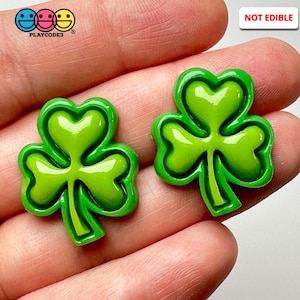 May include: Two green shamrock shaped resin embellishments with a glossy finish.  The shamrocks have a raised outline and a slightly darker green center.