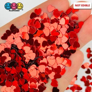 May include: A close-up of a hand holding a large amount of red heart-shaped glitter. The glitter is shiny and has a metallic finish. The text "NOT EDIBLE" is visible in the upper right corner of the image.