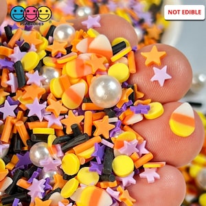 May include: A mix of Halloween sprinkles in orange, yellow, purple, and black. The sprinkles are shaped like stars, candy corn, and small circles. There are also white pearl-like sprinkles. The text "NOT EDIBLE" is visible in the image.