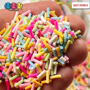 May include: A close-up of a handful of colorful sprinkles, including pink, yellow, blue, green, and white. The sprinkles are in the shape of long, thin cylinders. The text "NOT EDIBLE" is visible in the upper right corner of the image.