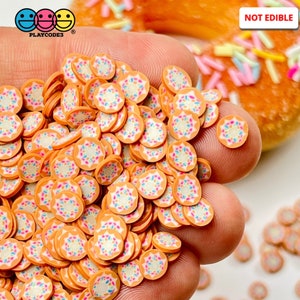 May include: A close-up of a hand holding a pile of small, round, orange and white donut-shaped sprinkles. The sprinkles are made of polymer clay and are not edible. The background shows a donut with colorful sprinkles.