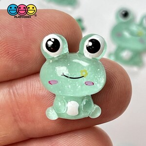 May include: A small, green, glitter-covered frog figurine with large black eyes and a white belly. The frog has a small yellow flower on its nose and pink cheeks.