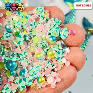 May include: A mix of green, blue, pink, and white resin sprinkles, beads, and charms. The sprinkles are shaped like flowers and seashells. The beads are pearl-like and iridescent. The charms are small and have a variety of shapes. The text "NOT EDIBLE" is visible in the image.