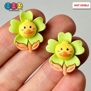 May include: Two green flower-shaped charms with a yellow duck face in the center. The flowers have orange stems and leaves. The charms are not edible. "NOT EDIBLE" is written in red on a white background.