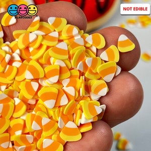May include: A close-up of a hand holding a pile of small, orange, yellow, and white candy corn shaped sprinkles. The sprinkles are not edible. The text "NOT EDIBLE" is visible in the upper right corner of the image.