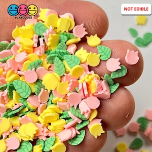 May include: A close-up of a hand holding a variety of colorful polymer clay sprinkles. The sprinkles are shaped like leaves, tulips, and other floral designs. The text "NOT EDIBLE" is visible in the upper right corner of the image.