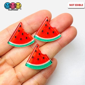 May include: Three watermelon slice-shaped charms with a bite taken out of each. The charms are red with black seeds and a green rind. The text "NOT EDIBLE" is in the upper right corner. The Playcode3 logo is in the upper left corner.